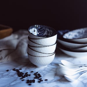 May include: A stack of four white ceramic bowls with a textured exterior and a black rim. The interior of the bowls features a blue and white floral design. Several white spoons with blue floral accents are also visible.