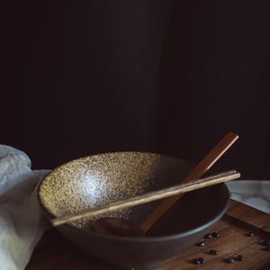 Pu&ograve; includere: Una ciotola in ceramica con interno maculato beige e marrone ed esterno marrone scuro. Un paio di bacchette di legno poggia sopra la ciotola e un cucchiaio di legno &egrave; posto all'interno. Piccoli fagioli neri sono sparsi attorno alla ciotola.