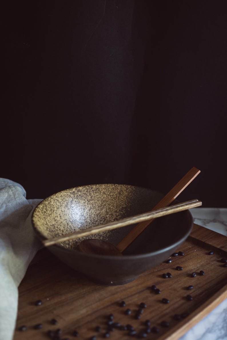 Pu&ograve; includere: Una ciotola in ceramica con interno maculato beige e marrone ed esterno marrone scuro. Un paio di bacchette di legno poggia sopra la ciotola e un cucchiaio di legno &egrave; posto all'interno. Piccoli fagioli neri sono sparsi attorno alla ciotola.