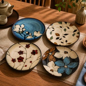 May include: A set of four round ceramic plates featuring distinct floral patterns. One plate has white flowers on a blue background, one has small red blossoms on a cream base, one has green and brown leaves on cream, and one has large blue and white flowers.
