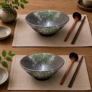 Pu&ograve; includere: Due ciotole in ceramica con smalto maculato verde e grigio scuro, ciascuna accompagnata da un cucchiaio di legno e un paio di bacchette in legno scuro. Le ciotole presentano un bordo largo e svasato e una finitura testurizzata su tovagliette beige.