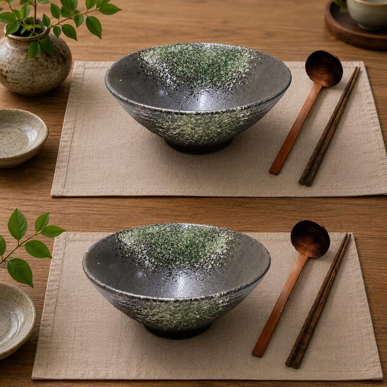 Pu&ograve; includere: Due ciotole in ceramica con smalto maculato verde e grigio scuro, ciascuna accompagnata da un cucchiaio di legno e un paio di bacchette in legno scuro. Le ciotole presentano un bordo largo e svasato e una finitura testurizzata su tovagliette beige.