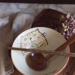 Könnte beinhalten: Keramikschüssel mit Katzenillustration, Holzlöffel und Essstäbchen. Die Schüssel hat einen rosa Rand und einen cremefarbenen Innenraum. Die Katze ist im Cartoon-Stil mit rosa Wangen gezeichnet. Auf einer Holzoberfläche.