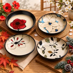 May include: A set of four round ceramic bowls with black rims. Each bowl features a unique hand-painted design: one with a large red poppy, one with white flowers on a blue background, one with a stylised white flower, and one with leafy vines.