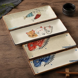 May include: A set of rectangular ceramic plates with hand-painted Japanese designs. The plates feature illustrations of fish, cats, and blue wave patterns. The plates have a cream background with brown trim.