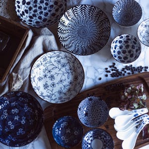 May include: An assortment of blue and white ceramic bowls and plates with various floral and geometric patterns. The collection includes serving bowls, small bowls, and spoons, arranged on a wooden tray and marble surface.