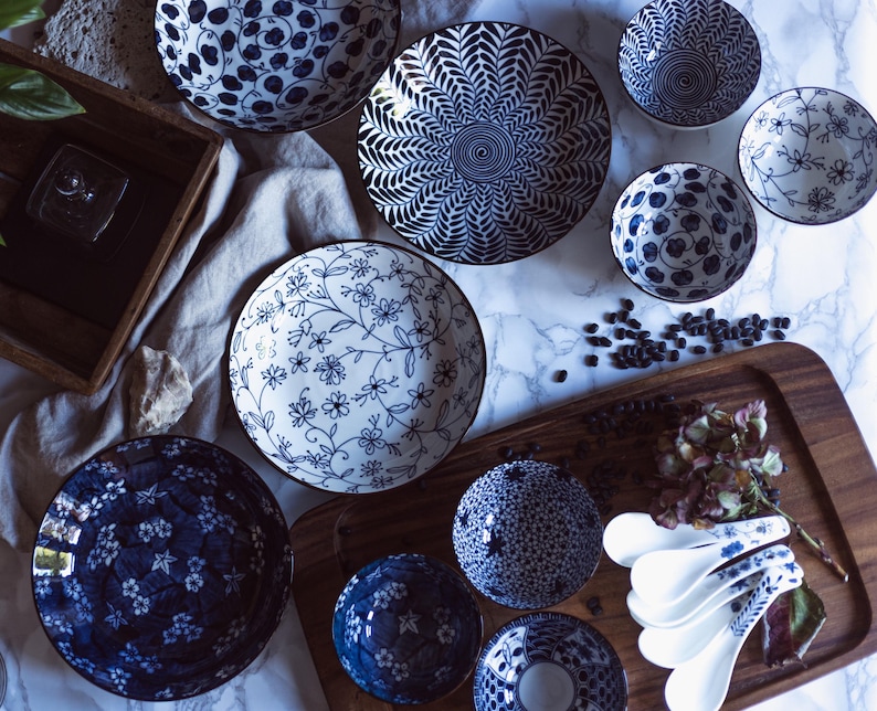 May include: An assortment of blue and white ceramic bowls and plates with various floral and geometric patterns. The collection includes serving bowls, small bowls, and spoons, arranged on a wooden tray and marble surface.