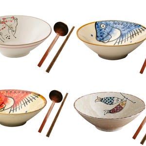 Könnte beinhalten: Vier Keramik-Schalen mit verschiedenen Designs, jede mit einem Holzlöffel und Essstäbchen. Die Schalen zeigen eine Katze, einen Fisch, einen roten Fisch und zwei Fische.