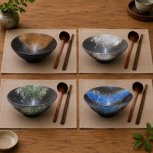 May include: Four ceramic bowls with a black base and a textured, speckled glaze in gold, white, green, and blue. Each bowl is accompanied by a dark wooden spoon and a pair of matching wooden chopsticks, all arranged on individual beige placemats.