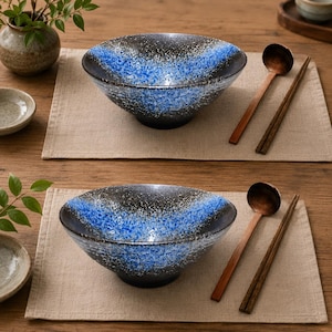 Pu&ograve; includere: Due ciotole in ceramica con esterno ed interno nero e un motivo a gradiente blu maculato. Ogni ciotola &egrave; accompagnata da un cucchiaio di legno e un paio di bacchette di legno scuro, disposti su tovagliette individuali in tessuto beige chiaro.