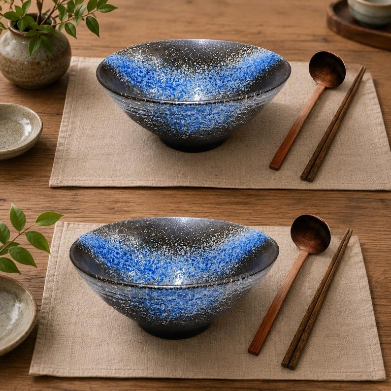 Pu&ograve; includere: Due ciotole in ceramica con esterno ed interno nero e un motivo a gradiente blu maculato. Ogni ciotola &egrave; accompagnata da un cucchiaio di legno e un paio di bacchette di legno scuro, disposti su tovagliette individuali in tessuto beige chiaro.