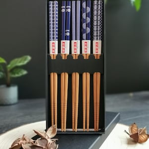 May include: A set of six wooden chopsticks in a black box. The chopsticks are brown and have a blue and white patterned design. The box has the text "竹製箸" in Japanese.
