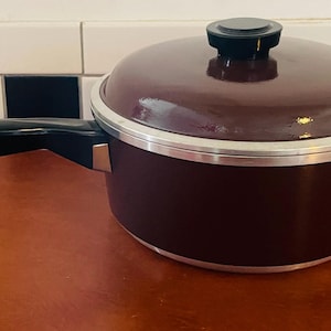 May include: A maroon saucepan with a matching lid and a black handle. The pot has a silver rim and a black knob on the lid. The handle extends to the left, and the pot sits on a wooden surface.