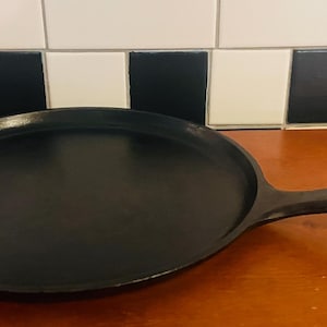 May include: A black cast iron griddle pan with a long handle. The pan is round with a flat cooking surface. The handle has a hole at the end for hanging. The background features a black and white tiled wall.