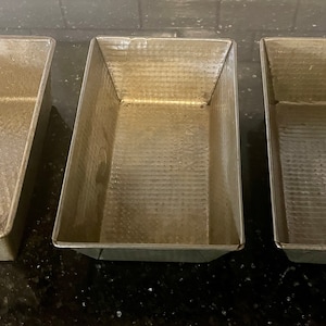 Puede incluir: Tres moldes para pan de plata, todos del mismo tamaño y forma, están alineados en una encimera negra. Los moldes son de metal y tienen una superficie texturizada.