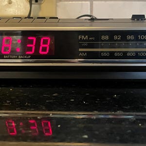 May include: A General Electric AM/FM clock radio with a silver and black rectangular housing. The front features a digital red LED display showing 8:38, an analog radio tuning dial with frequency scales, and the General Electric logo on the left.