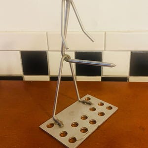 May include: A handcrafted metal sculpture of a person, made from nuts, bolts, and wire. The figure is standing on a rectangular base with several circular cutouts. The sculpture has a unique, industrial aesthetic.