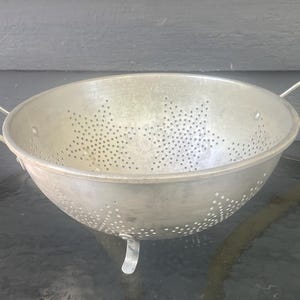 Puede incluir: Un colador redondo de metal color plata con dos asas laterales y tres patas pequeñas. El cuenco del colador está perforado con numerosos agujeros pequeños dispuestos en un patrón de estrella en la superficie interior y en la base.