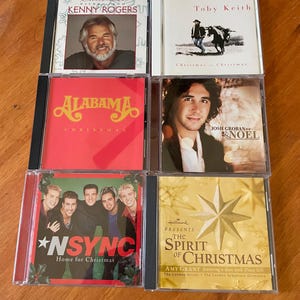 May include: A collection of six Christmas music CDs. Featured artists include Kenny Rogers, Toby Keith, Alabama, Josh Groban, *NSYNC, and Amy Grant. The album covers display various images and text, including artist names and album titles.