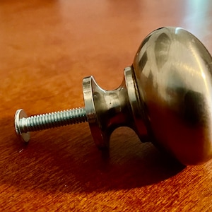 Może przedstawiać: Srebrny gałka do szafki o okrągłym, lekko owalnym kształcie. Gałka ma śrubę przymocowaną do tyłu.