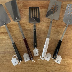 Può includere: Una collezione di cinque spatole in metallo con vari design e forme del manico. Ogni spatola presenta una lama unica, incluse opzioni piene e fessurate. I manici sono fissati con rivetti e sono dotati di etichette bianche numerate.