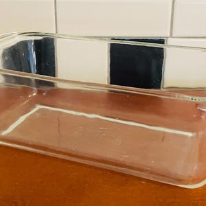 Op de afbeelding: Een heldere glazen cakevorm met een rechthoekige vorm en afgeronde hoeken. De vorm heeft aan elk uiteinde een handvat en staat op een bruin oppervlak. De achtergrond heeft een zwart-wit geblokt patroon.
