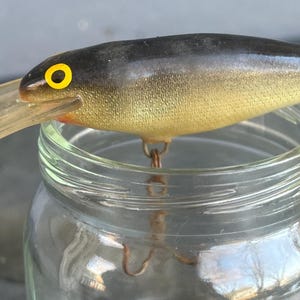 May include: A vintage fishing lure with a black and yellow body, a clear plastic bill, and a treble hook. The lure has a yellow eye with a black pupil. The lure is resting on a glass jar.
