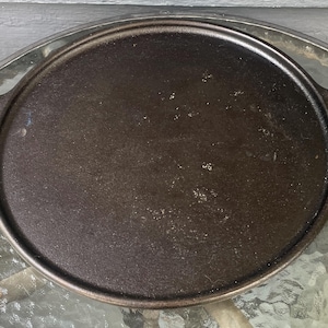 May include: A round, black cast iron griddle featuring a flat cooking surface with a slightly raised outer rim. The griddle includes two integrated, rectangular-shaped handles on opposite sides, designed for a secure grip during use.
