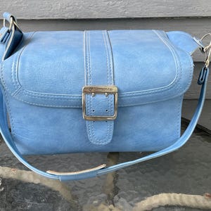 Könnte beinhalten: Eine hellblaue Umhängetasche mit glatter Oberfläche, die über einen mittigen vertikalen Riemen mit silberfarbener Metallschnalle verfügt. Die Tasche hat einen verstellbaren Schulterriemen mit silberfarbenen Beschlägen und ein Samsonite-Logo.