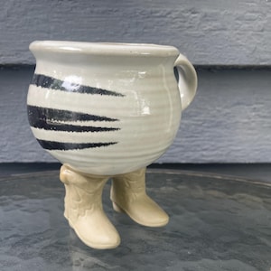 May include: A ceramic mug with a rounded body and a handle, featuring a cream-colored glaze with horizontal black brushstroke accents. The mug is supported by two sculpted beige ceramic cowboy boots that serve as the base of the vessel.