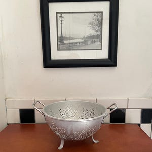 May include: A silver metal colander with small holes and three short legs sits on a wooden surface. The colander has two handles and is positioned below a framed black and white photograph of a street scene.