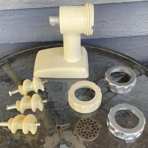 May include: A vintage cream-coloured meat grinder with various components, including a base, augers, and grinding plates. The grinder features a cream-coloured body and several metal parts. The components are arranged on a glass surface.