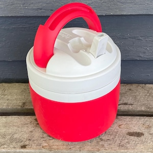 Puede incluir: Una jarra de agua Igloo roja y blanca con un asa roja. La jarra tiene una tapa blanca con una boquilla abatible. El cuerpo de la jarra es rojo y el asa está arqueada para facilitar su transporte. La jarra está diseñada para mantener las bebidas frías.