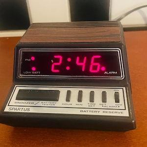 Puede incluir: Un reloj despertador digital Spartus vintage que muestra las 2:46 PM en dígitos de color rosa brillante. El reloj tiene una parte superior con vetas de madera, una cara negra y varios botones de función, incluyendo repetición y ajuste de alarma. Se ve el texto "Battery Reserve".