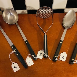 May include: A collection of vintage kitchen utensils, including two ladles, two spoons, and a potato masher. The utensils have stainless steel heads and black handles. Each utensil has a tag attached with a number.