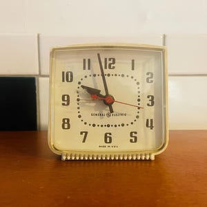Könnte beinhalten: Ein Vintage-Wecker von General Electric mit cremefarbenem Zifferblatt und quadratischer Form. Die Uhr hat schwarze Ziffern, einen roten Sekundenzeiger und den Text "MADE IN USA". Die Uhr steht auf einer Holzoberfläche.