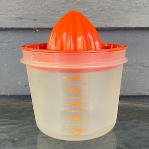 May include: An orange citrus juicer with a clear plastic measuring cup. The juicer has a ribbed, cone-shaped reamer and a rim that fits over the cup. The cup has orange measurement markings from 100 ml to 500 ml.