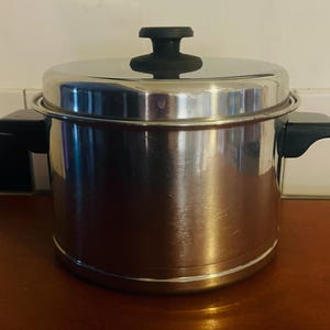 May include: A stainless steel cooking pot with a lid and black handles. The pot is sitting on a wooden surface. The lid has a black handle. The pot is designed for cooking.