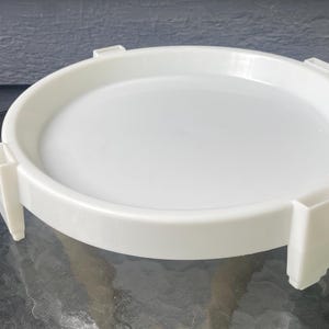 May include: A circular white plastic stand featuring a flat, recessed center platform surrounded by a raised rim. The structure is supported by four evenly spaced, rectangular vertical legs that extend downward from the outer edge of the frame.