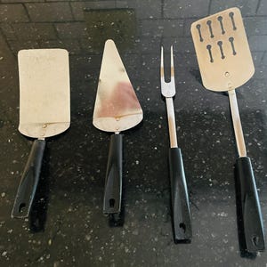 May include: Four stainless steel kitchen utensils with black handles. The set includes a rectangular spatula, a pie server, a two-pronged fork, and a slotted spatula. The utensils are arranged on a dark surface.