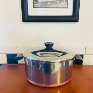 May include: A stainless steel pot with a copper-colored base and a black handle and lid. The pot is sitting on a wooden surface. A framed black and white picture hangs in the background.