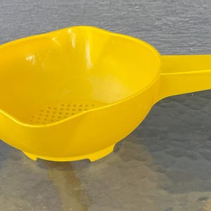 May include: A bright yellow plastic colander with a rounded bowl shape, a long flat handle featuring a small circular hole at the end, and a series of small drainage holes located at the bottom centre of the basin for straining liquids.