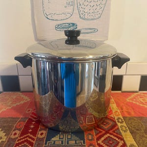 Può includere: Una pentola da cucina in acciaio inossidabile lucido con coperchio e manici neri. La pentola è molto riflettente. Il coperchio ha un pomello nero. La pentola è su una tovaglia fantasia colorata.