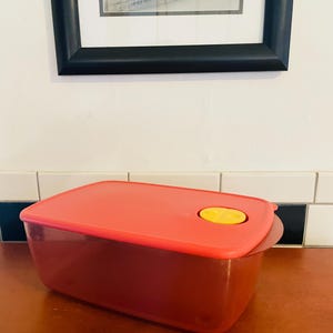 Könnte beinhalten: Ein roter, rechteckiger Lebensmittelbehälter mit passendem Deckel. Der Deckel hat einen gelben, kreisförmigen Knopf. Der Behälter steht auf einer Holzoberfläche, mit einem gerahmten Bild im Hintergrund.