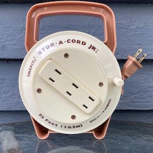 May include: A vintage Stor-A-Cord Jr. cord reel in cream and peach. The reel has a handle and a built-in outlet with three sockets. The text "25 Feet (7.62m)" is printed on the reel, along with the words "Snapit" and "Wind/Unwind".