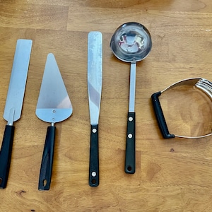 Puede incluir: Cinco utensilios de cocina con mangos negros sobre una superficie de madera. Los utensilios incluyen un servidor de pastel, una espátula, un cortador de masa, un cucharón y una espátula de borde recto.