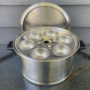 May include: A stainless steel egg poacher pan with a matching lid and black side handles. The insert features six individual circular cups for poaching eggs. The side of the pan is engraved with the brand name Lifetime and stainless steel.