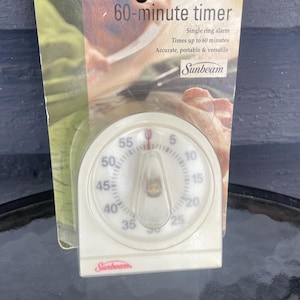 以下が含まれることがあります： 赤色の針が付いた白い60分タイマー。緑とベージュの背景にパッケージされています。タイマーは単一のリングアラームを備え、正確でポータブルで用途が広いと表示されています。「Sunbeam」のブランド名が見えます。