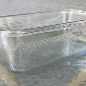 Op de afbeelding: Een rechthoekige broodvorm van helder glas met een decoratief reliëfpatroon langs de bovenrand. De schaal heeft gladde, afgeronde hoeken en een transparante afwerking, wat een klassiek en functioneel ontwerp laat zien.