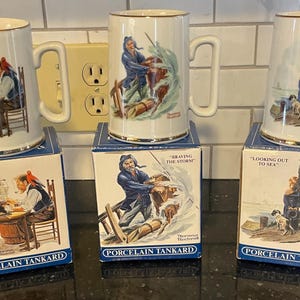 May include: Three porcelain tankards with gold rims, each featuring a different nautical scene. The tankards are displayed on their original boxes, which also depict the scenes. The boxes have text that reads "For a Good Boy", "Braving the Storm", and "Looking Out to Sea".